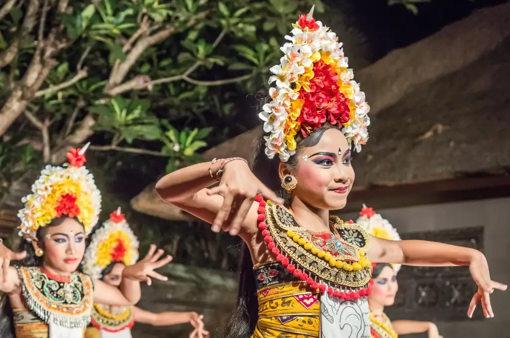 A group of five performers are captured during an apparent stage performance. The central figure is a young woman in ornate traditional dress featuring geometric patterns and vibrant hues of yellow, red, and black, complemented by elaborate gold jewelry and bracelets. Her dark hair is adorned with a striking headdress of flowers in shades of red, white, and yellow, and her face is painted with colorful stage makeup. The woman's pose is theatrical, with her hands gracefully extended, suggesting a moment of dance or ritual. This photograph would be fitting for use in projects relating to global travel, art, performance, tradition, and cultural celebrations.