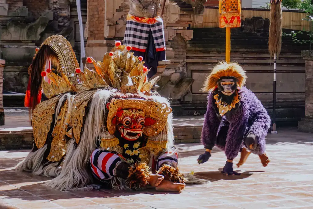 Two costumed performers pose in a paved square in Indonesia. One performer is crouched with feet visible wearing a costume with red and white stripes and a head piece with a red face. The other performer is wearing a purple furry costume and has an animal mask on the face. This photo would be appropriate for projects needing imagery depicting culture, performance, and travel.