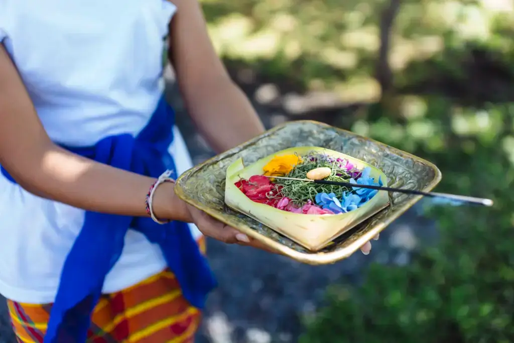 A child holds a gold square platter containing a woven leaf offering in an outdoor setting. The woven offering contains colorful flower petals and what looks like a small, lit incense stick. The child wears a white top, bright blue sash, and a patterned orange, red, and yellow wrap around the waist. Soft, natural lighting creates a calm and peaceful mood. This photo could be used for projects centered on religious and spiritual practices, travel, or cultural celebrations.
