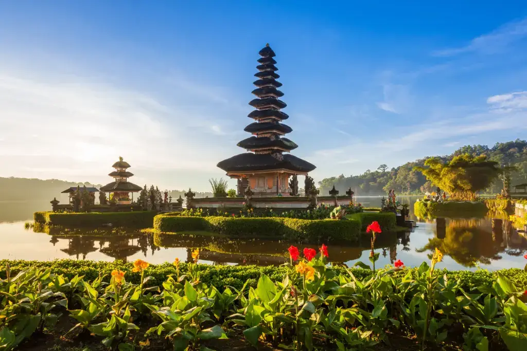 This tranquil photograph showcases a temple complex nestled on the edge of a serene lake. The still water reflects the intricate architecture of the structures, which include a multi-tiered pagoda. Lush greenery and vibrant flowers frame the foreground, enhancing the natural beauty of the scene. The time of day appears to be daytime with golden hour light reflecting on the trees and water, suggesting early morning light. This striking image is ideal for projects focusing on travel, tourism, spirituality, or Southeast Asian culture. Its peaceful and scenic nature could serve as a visual metaphor for harmony and natural beauty.