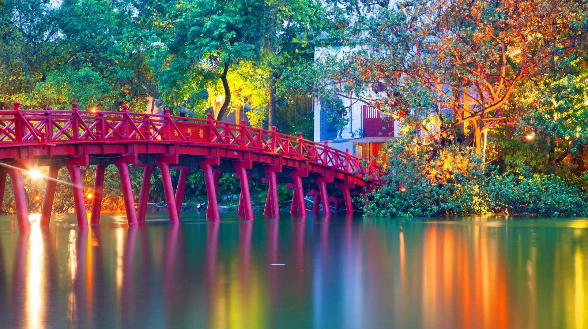 This beautiful photograph features a traditional-style red bridge illuminated by nearby lights, reflected in the still water. The scene is set among lush greenery, including mature trees, with a building partially visible in the background. The lighting suggests evening, adding a tranquil mood. This idyllic location could be used for travel and tourism projects, showcasing culture and architecture, or promoting a sense of peaceful escape.
