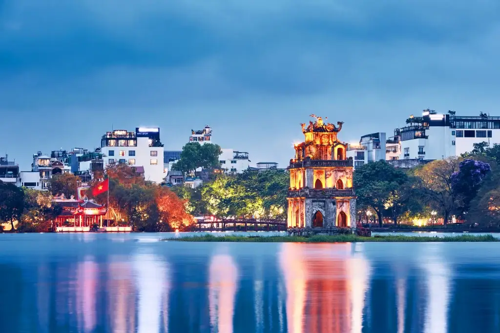 This scenic photo captures a tranquil cityscape featuring the illuminated Turtle Tower on an islet in Hoan Kiem Lake, Hanoi. The water reflects the tower's warm light and the lights of the surrounding buildings. This image is suitable for projects related to travel, architecture, Vietnamese culture, or tranquil urban scenes. The dark sky evokes a peaceful mood.