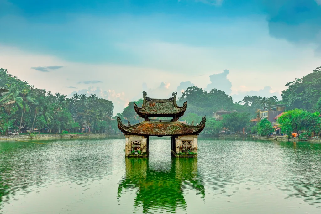 An ancient, two-tiered temple gateway stands in the middle of a tranquil lake. The temple structure features elaborate designs and a weathered appearance, with reddish-brown roofing and light-colored supports. Lush green trees line the shores of the lake, and rolling hills fade into a light gray sky. The still water reflects the temple, creating a mirror image, enhancing the sense of peace. This idyllic scene evokes a sense of history and spirituality, perfect for travel brochures, cultural documentaries, and serene nature scenes.