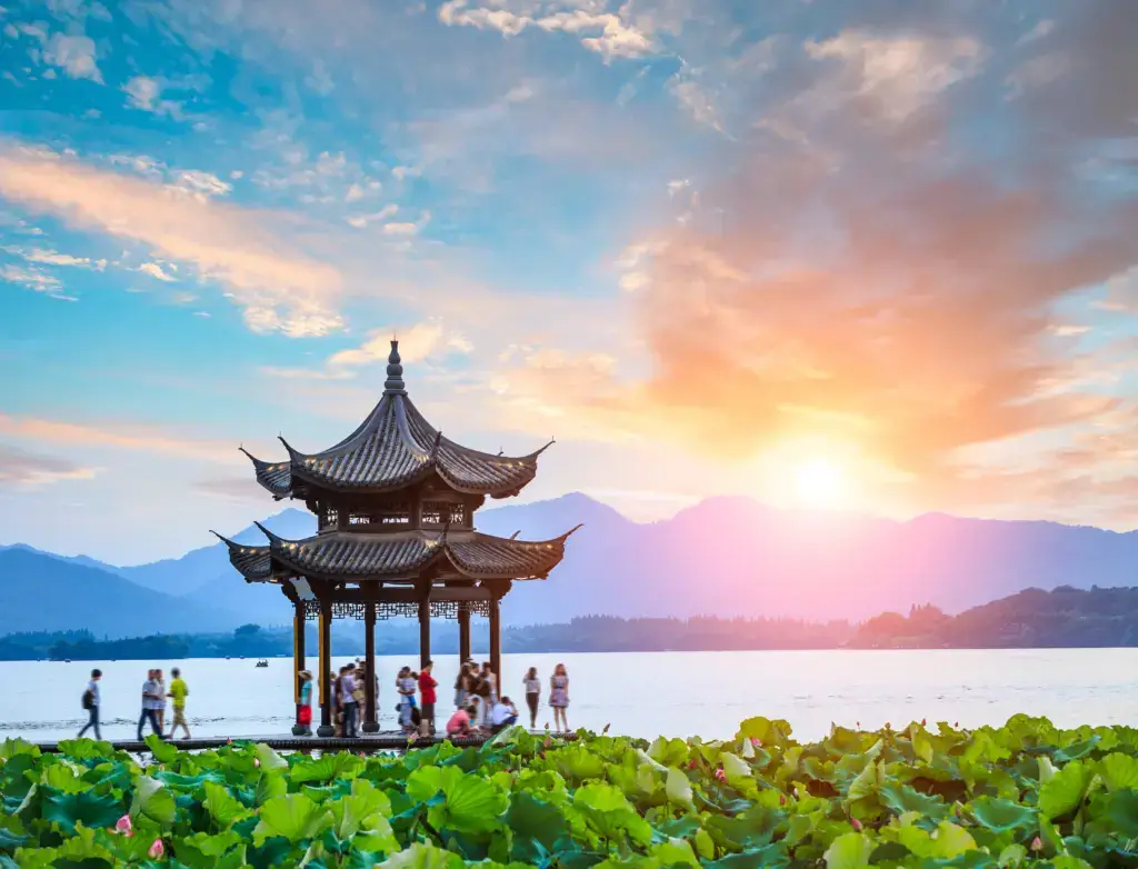This beautiful photograph captures a pagoda near West Lake in China at sunrise or sunset. A group of people stroll along the waterfront. Lush vegetation surrounds the pagoda and walkway. The mountains in the background provide a scenic vista. The photograph would be useful for travel and tourism projects related to the Chinese landscape, architecture, and culture, as well as for themes of peace, beauty, and harmony with nature. The image is well-composed, with vibrant colors and lighting.