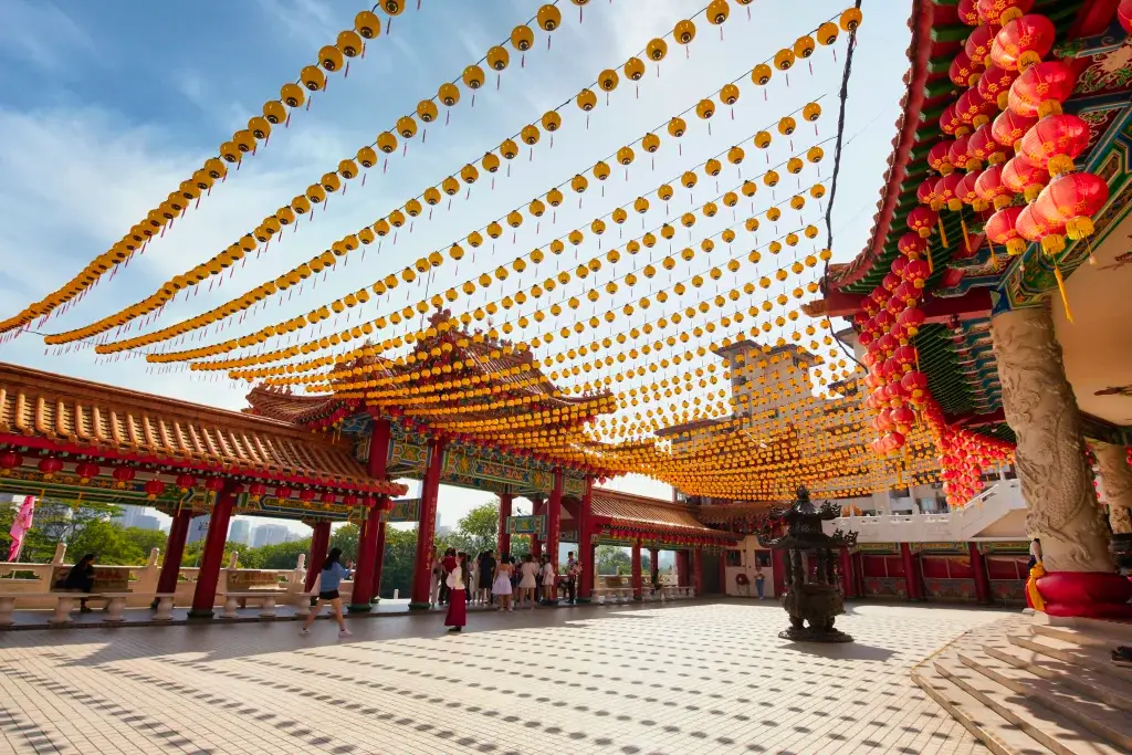 A bright and expansive wide-angle shot features hundreds of yellow and red paper lanterns strung in rows across the open courtyard of a traditional temple. Several people, including a woman in a blue shirt and another in a red skirt, walk across the light-colored tiled ground, which is decorated with long, linear shadows cast by the decorations above. The ornate temple structures with tiered roofs and intricate carvings stand under a clear blue sky, while distant urban buildings are visible on the horizon. This festive and culturally evocative photo is perfect for themes related to travel, spirituality, cultural heritage, and seasonal celebrations like the Lunar New Year.